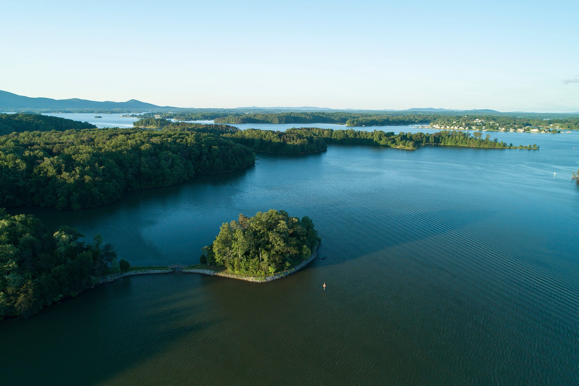 An island sits in calm blue waters, surrounded by lush green trees, with mountains visible on the horizon under a clear sky.
