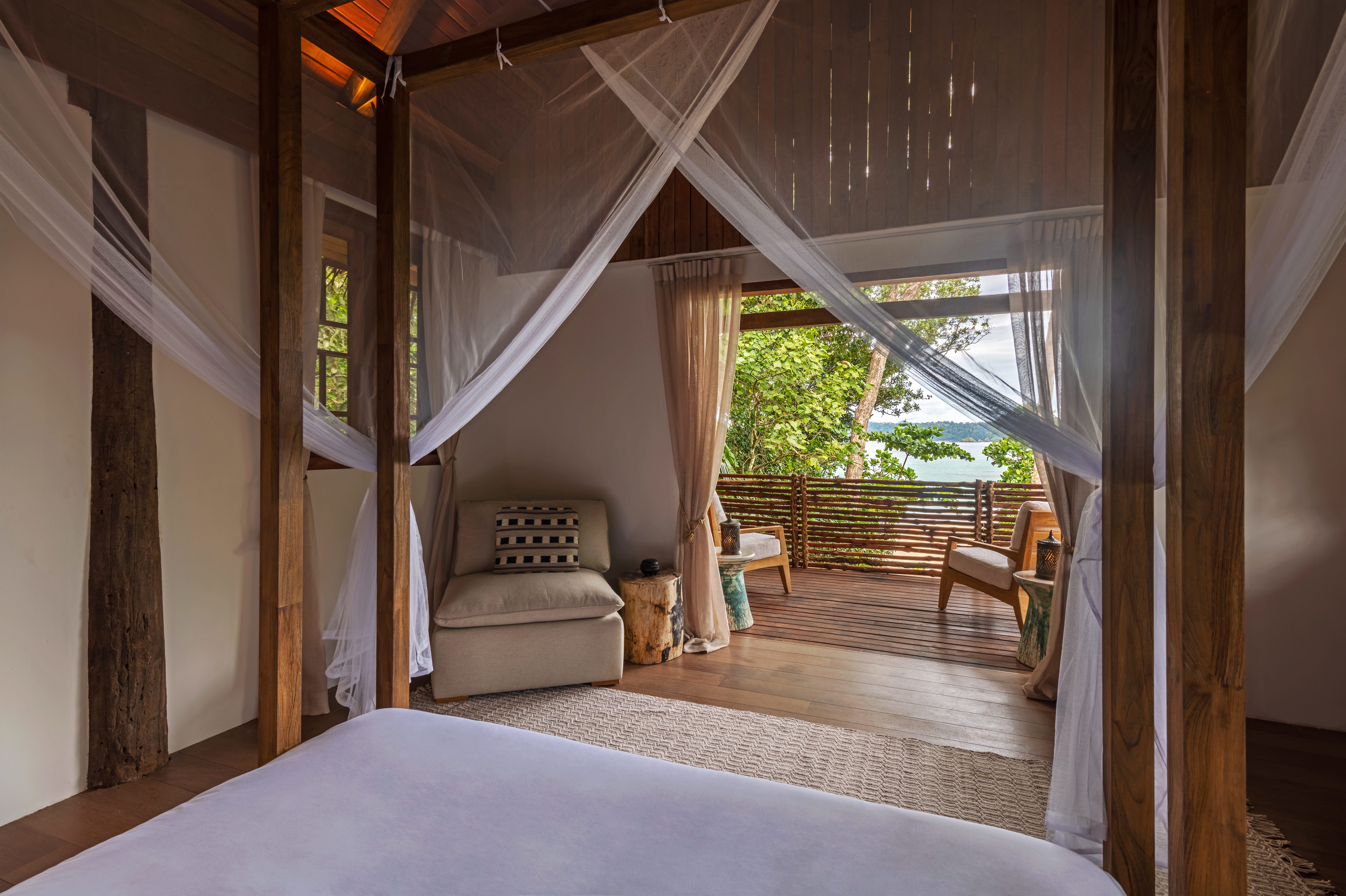 A bed with mosquito netting beside a cozy deck and seating area overlooks lush greenery and water.