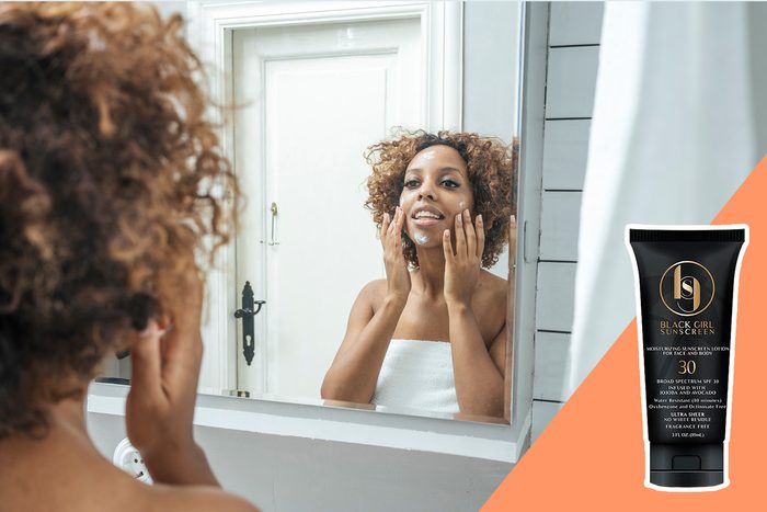 Woman applies sunscreen cream while looking in mirror; bathroom setting. Text on bottle: 