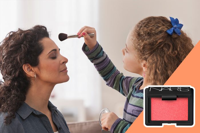 Child applies makeup on woman's face; woman sits with eyes closed. Blush product shown in corner. Bright, indoor setting with soft light.