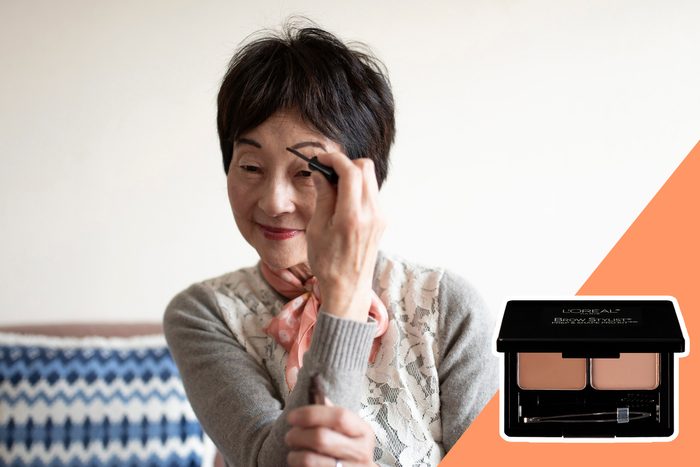A woman applies eyebrow pencil, smiling in a room with a patterned pillow. An inset shows a Lâ€™OrÃ©al brow kit on an orange background.