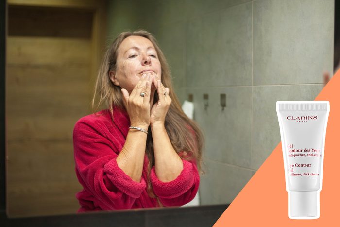 A person applies cream to their face while looking in a bathroom mirror. A Clarins Eye Contour Gel tube is displayed beside them.