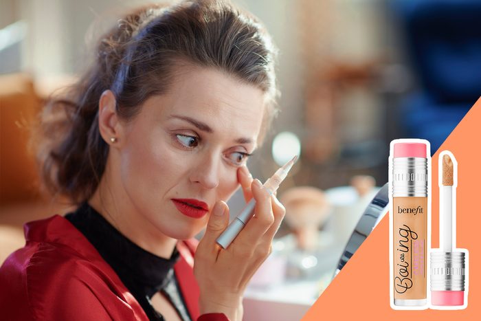 Woman applies concealer pen under eyes, sitting in a well-lit room with makeup products nearby. Image features Benefit Boi-ing Cakeless Concealer.