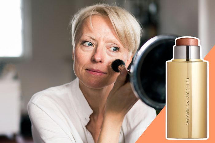 A person applying makeup with a brush while looking in a mirror, next to a gold tube labeled 