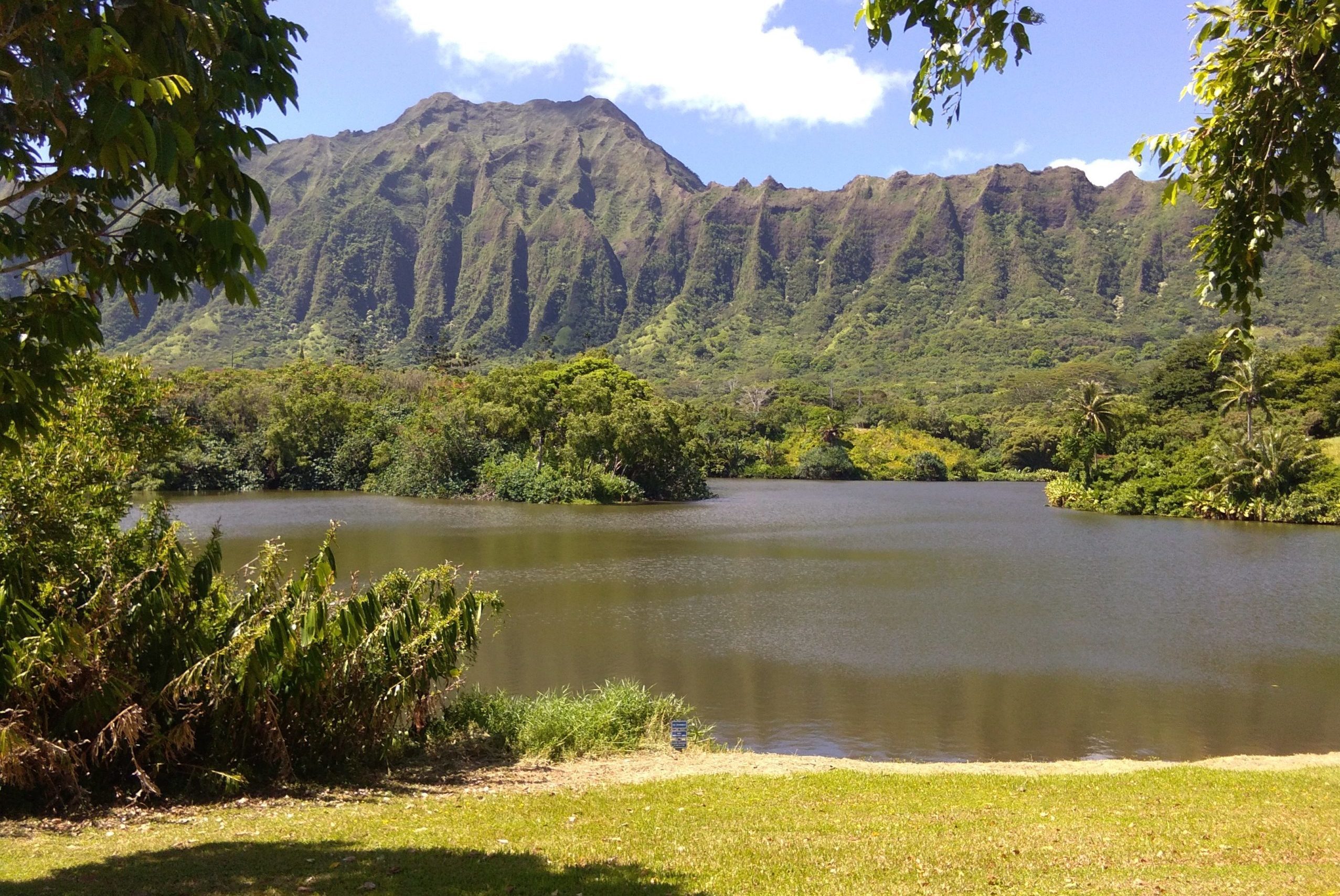 Mountains rise behind a calm lake, surrounded by lush greenery and trees under a partly cloudy sky.