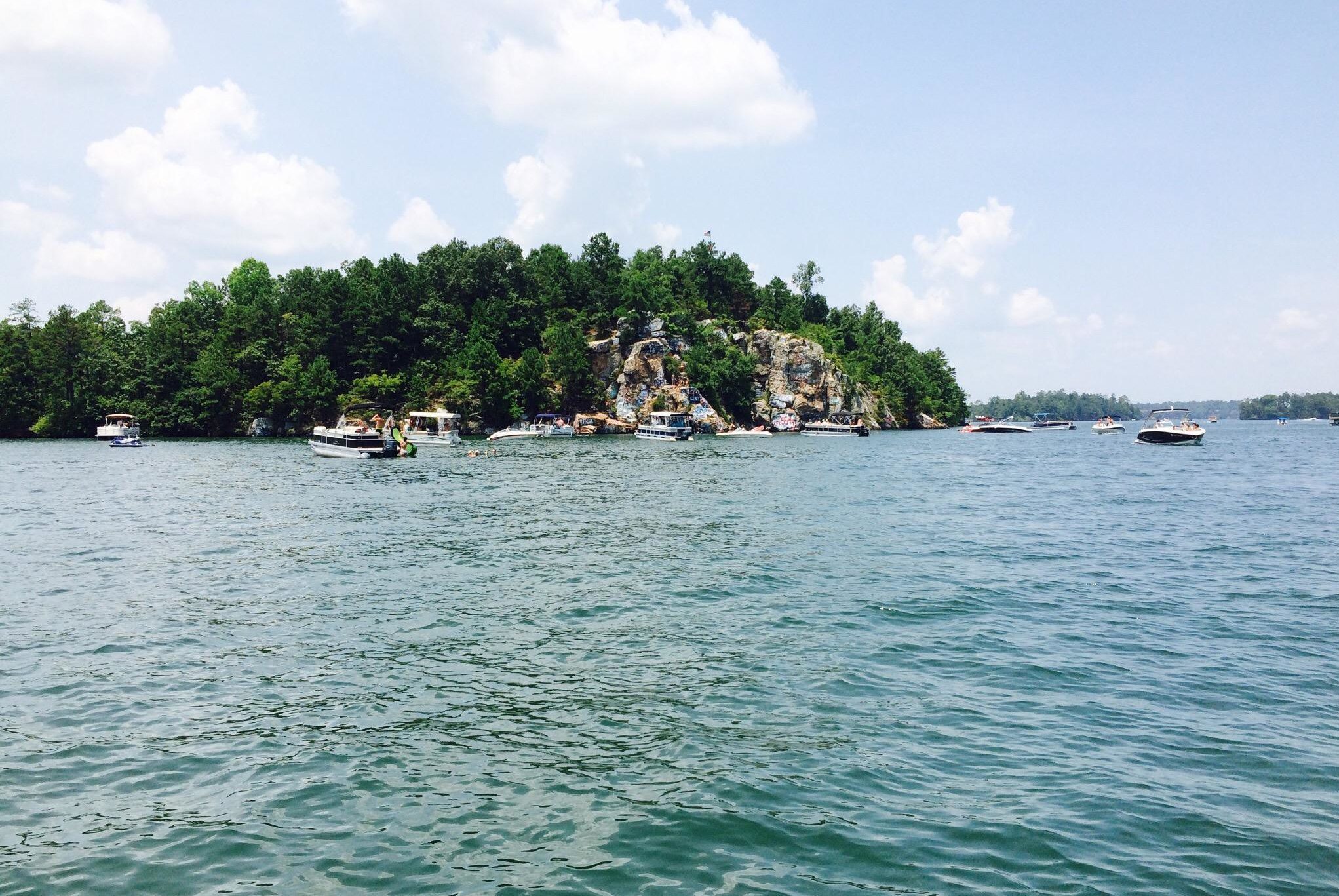 Boats float near a tree-covered rocky island, with people swimming nearby in a calm, expansive lake under a partly cloudy sky.