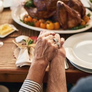 Hands hold each other across a table set with dishes, napkins, a roasted turkey, and wine, suggesting a festive meal gathering.