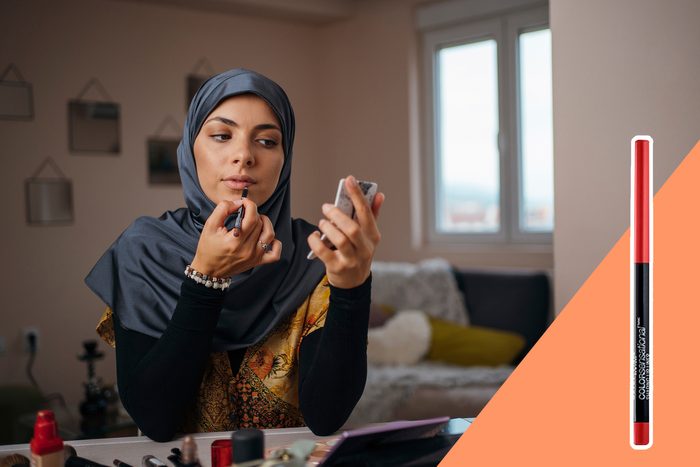 A person applies lipstick while holding a mirror, sitting in a cozy room with wall decorations and a window.