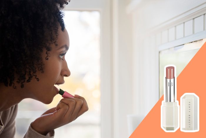 Person applying lipstick, facing a window, with a Fenty Beauty lipstick tube shown on an orange overlay.