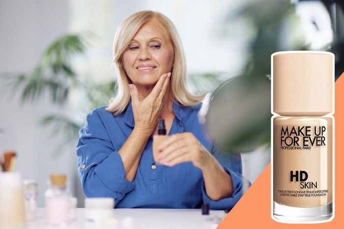 A woman applies liquid foundation to her face at a table with various cosmetics. A bottle labeled 