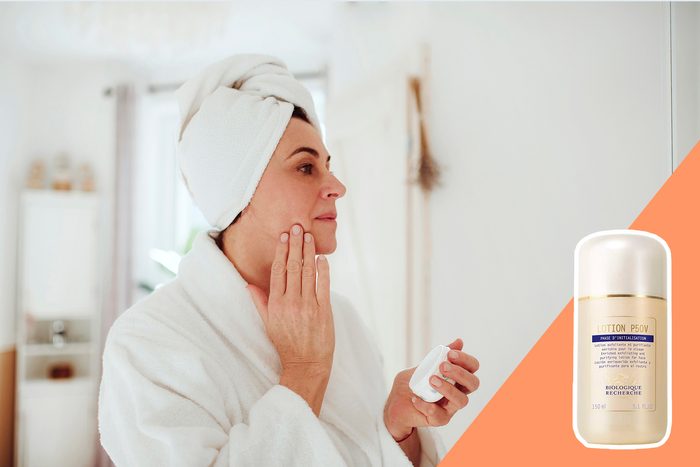 Person applies lotion to face, standing in a bright bathroom, towel wrapped on head. Bottle of 