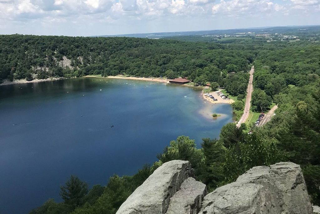 A serene lake reflects the sky, surrounded by dense forest and a distant road. People relax by the beach near a pavilion.