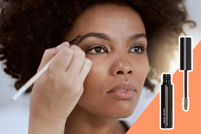 Woman applies eyebrow makeup with brush; black makeup tube labeled 