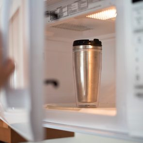 A metallic travel mug sits inside an open microwave, with a hand reaching to close the door, in a kitchen setting.