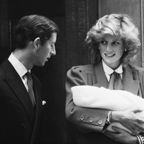 Woman smiling while cradling a baby, standing beside a man in a suit, outside a building entrance. Black and white photograph.