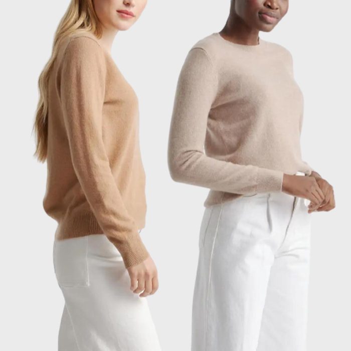 Two people wearing beige sweaters and white pants stand against a plain background.
