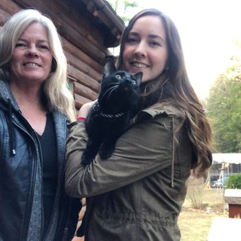 A black cat held by a smiling woman stands next to another woman, outside near a wooden house surrounded by trees.