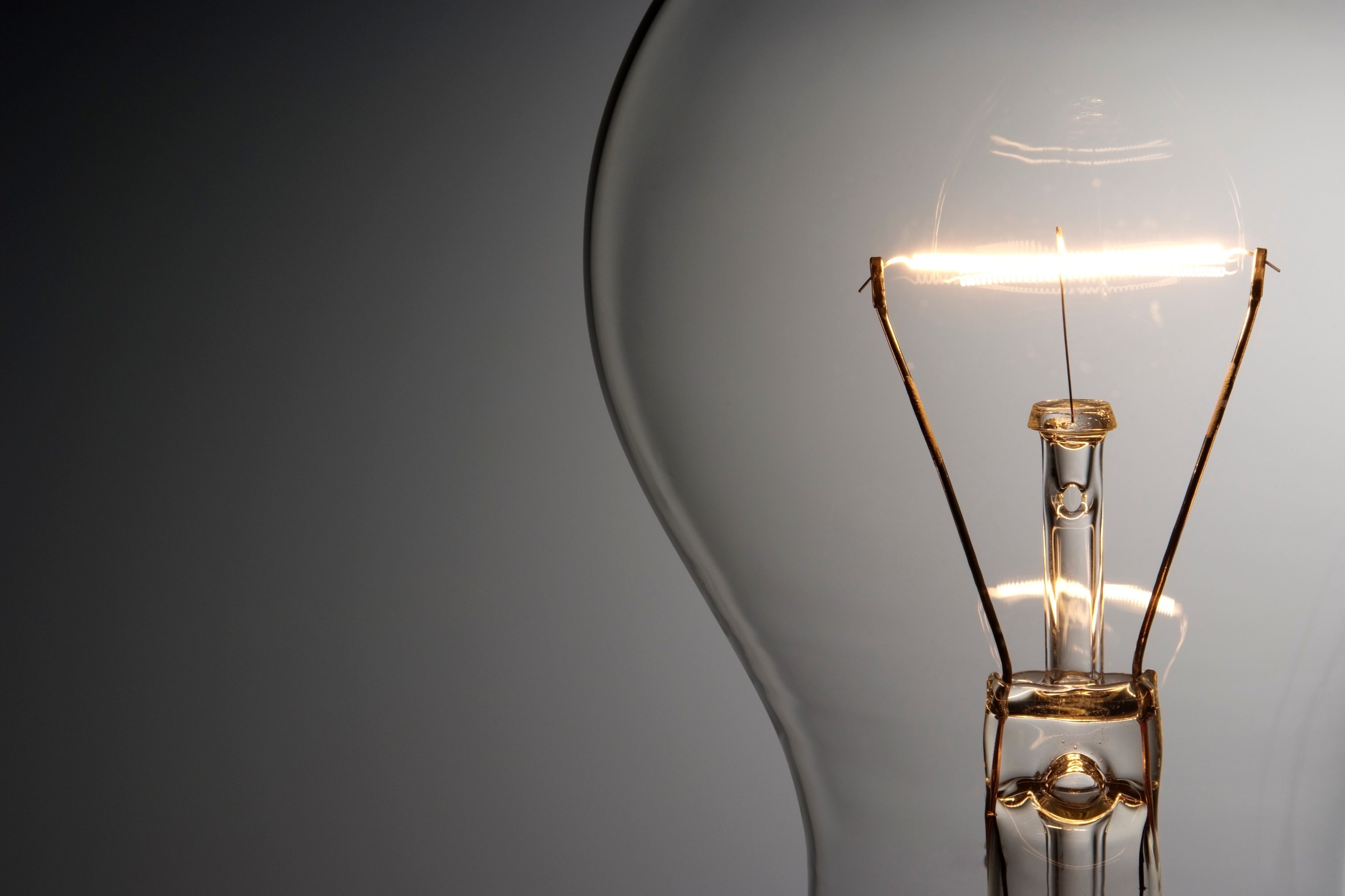 A light bulb filament glowing against a plain, dark background, illuminating the close-up glass encasement.