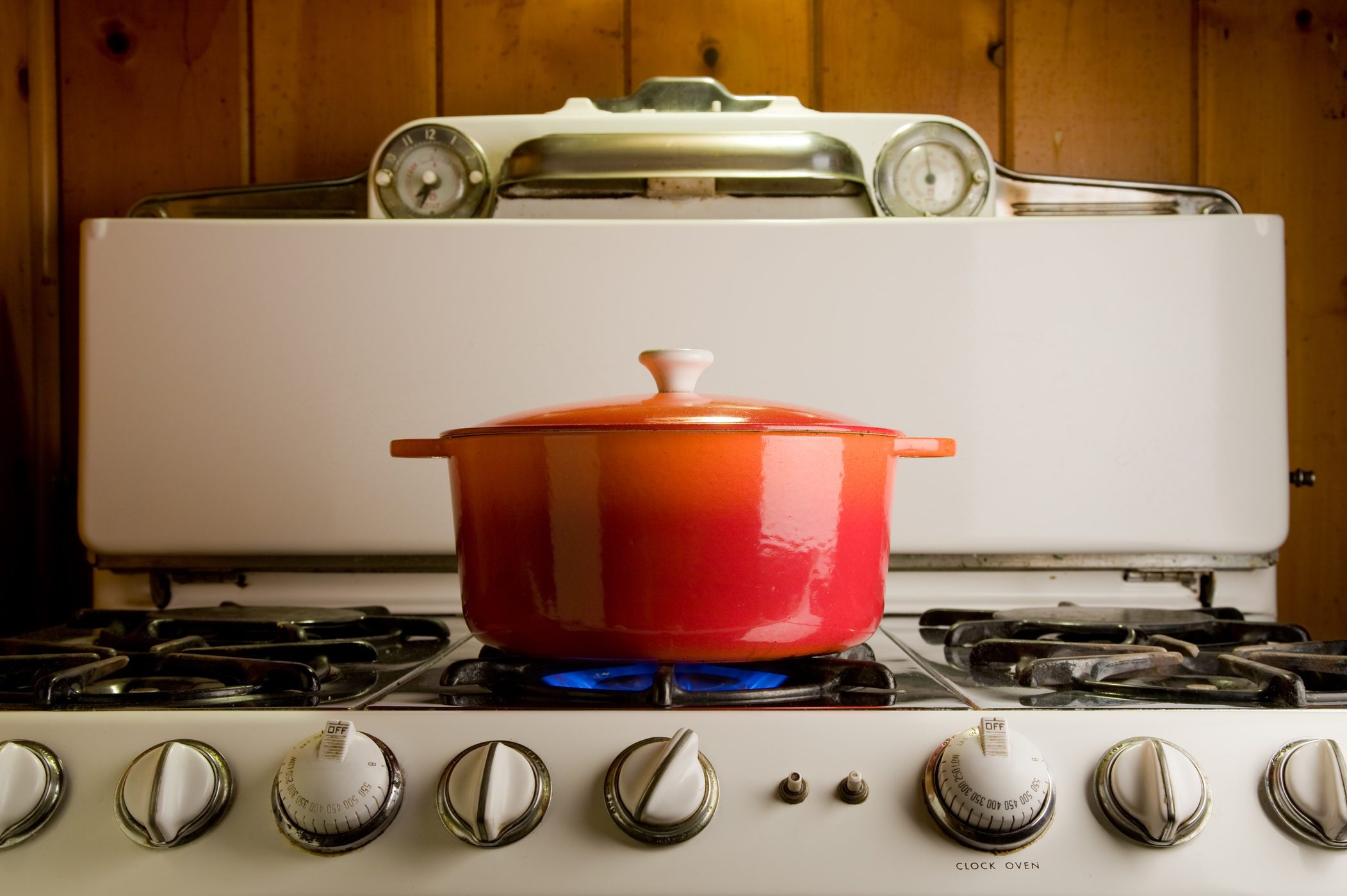 Red pot simmers on stove, surrounded by wooden kitchen backdrop, with blue flame underneath.