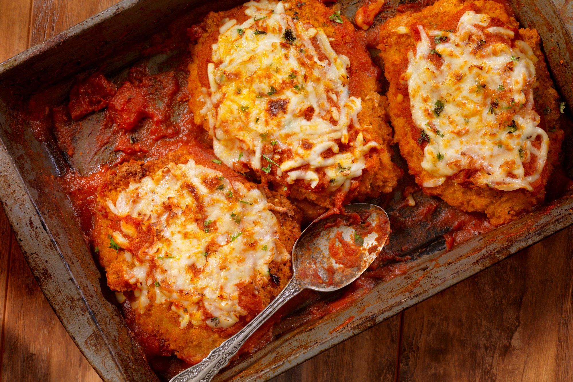 Baked chicken Parmesan topped with melted cheese sits in a rustic metal pan, surrounded by tomato sauce, with a spoon resting nearby.