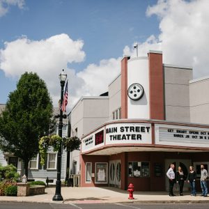 Theater marquee displays show dates; three people nearby chat. American flag waves beside lamp post. Sunny day with clouds above, surrounded by tree and plants.
