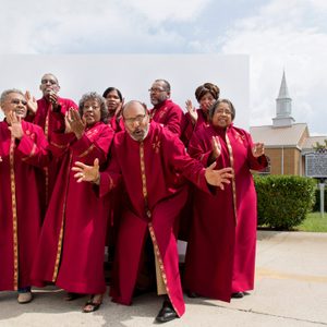 Choir members in red robes singing energetically outside with a church and sign in the background under a cloudy sky.