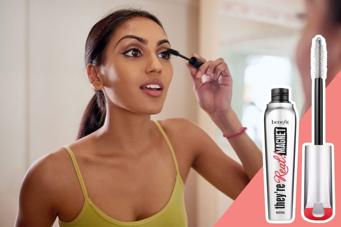 Woman applies mascara while looking in mirror, wearing a green top. Text: 