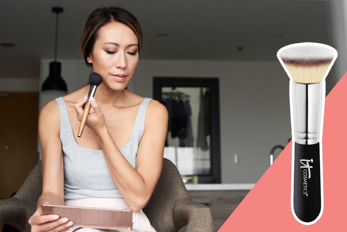 Woman applies makeup with a brush, sitting in a modern room. An inset image shows an 