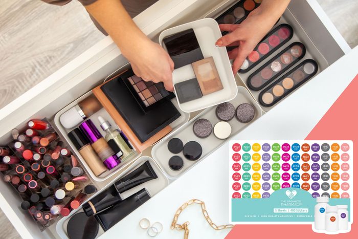 Hands organize cosmetics in a drawer with lipsticks, palettes, and bottles. A sheet of colorful, date-labeled stickers lies nearby.