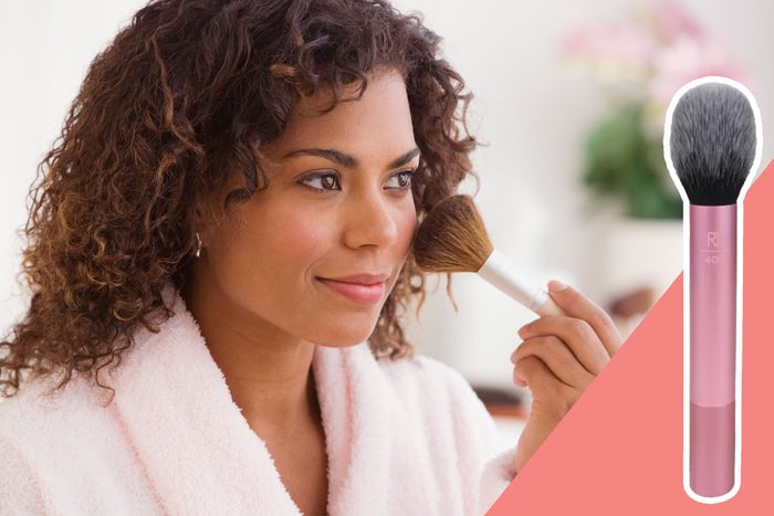 Woman applies makeup with a brush, wearing a pink robe, in a softly lit room. A close-up of a pink-handled brush is shown.