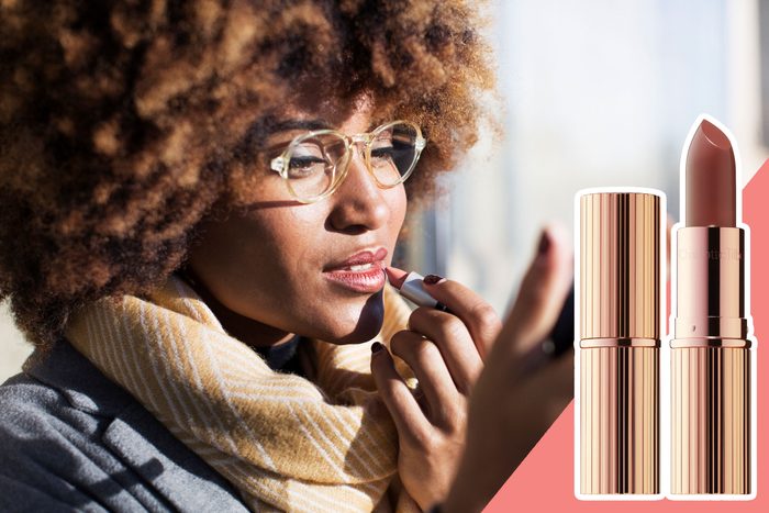 Person applies lipstick while looking in a mirror, wearing glasses and a scarf, with a lipstick tube displayed alongside in soft daylight.