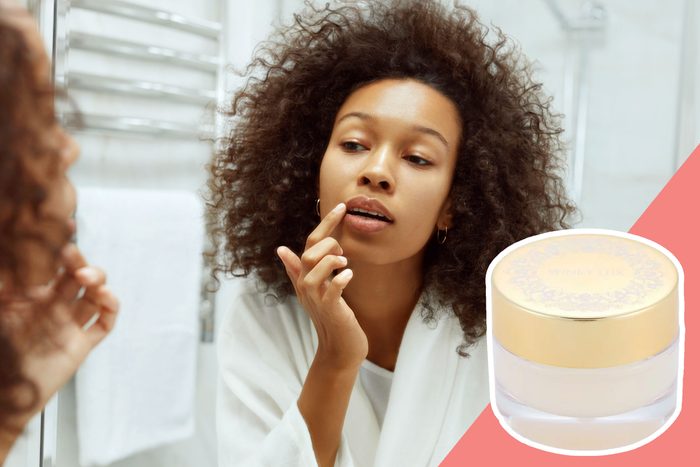 Person applies lip balm in a bathroom, wearing a white robe, near a wall-mounted towel. A jar labeled 