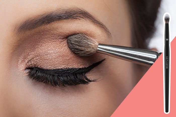 Eye receives makeup application with a brush, showing metallic eyeshadow and eyeliner. Right corner displays Sephora brush on a pink background.