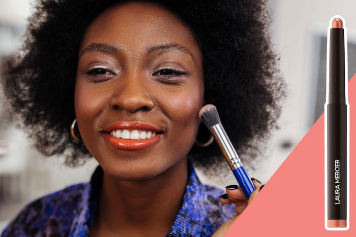 Woman smiles while applying makeup with a brush; a Laura Mercier cosmetic stick is displayed on the side.
