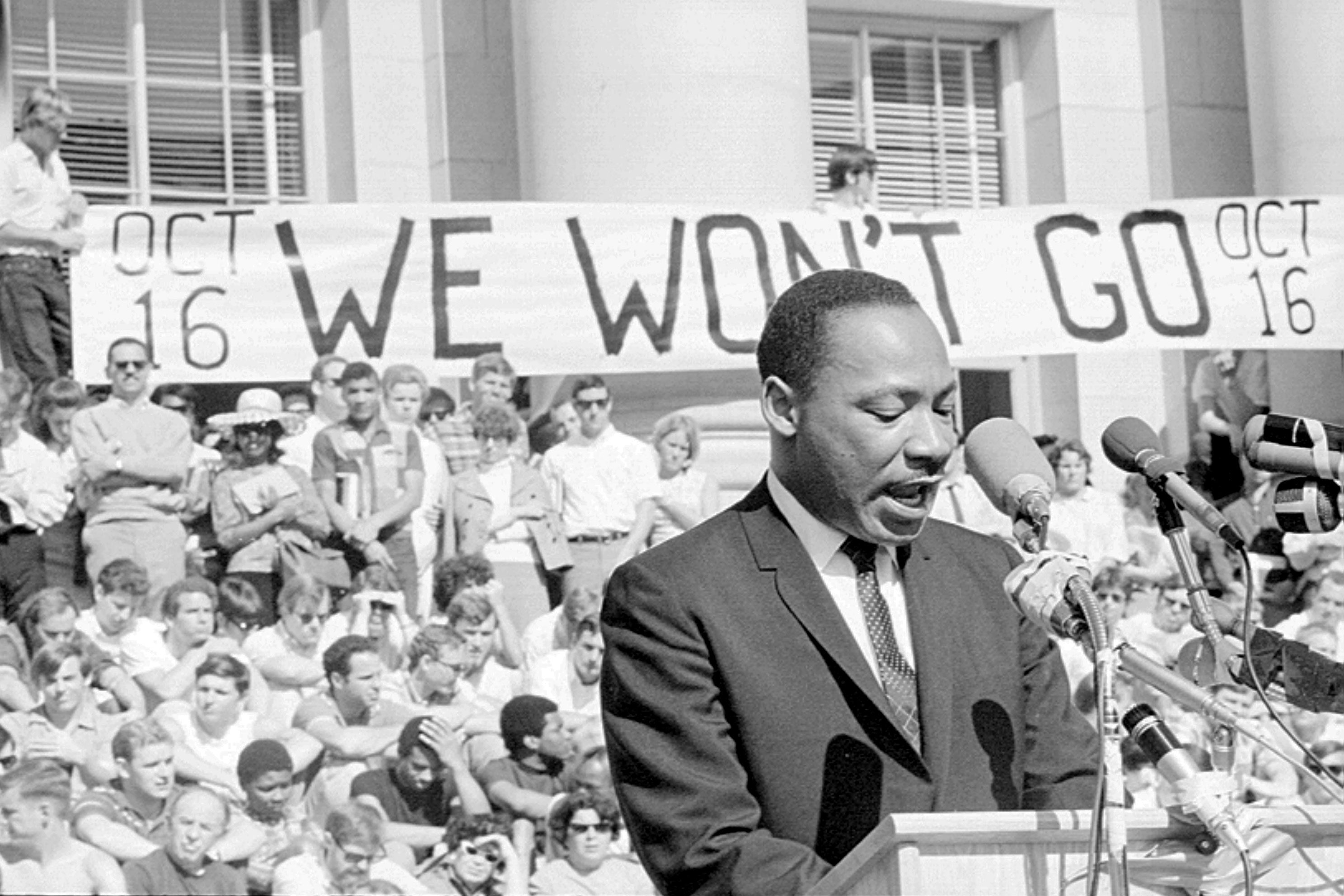 A man speaks at a podium; crowd holds a "WE WON'T GO" banner outside a building during a protest.
