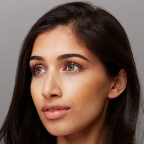A woman's face looks upward, showing detailed makeup and long lashes, against a plain gray background.