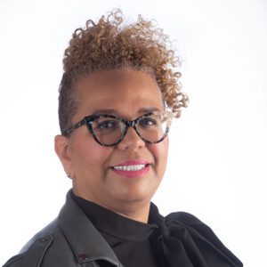 A person with curly hair and glasses smiles slightly, wearing a dark top, against a plain white background.