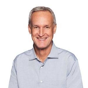 A person smiles, wearing a light blue button-up shirt against a plain white background.