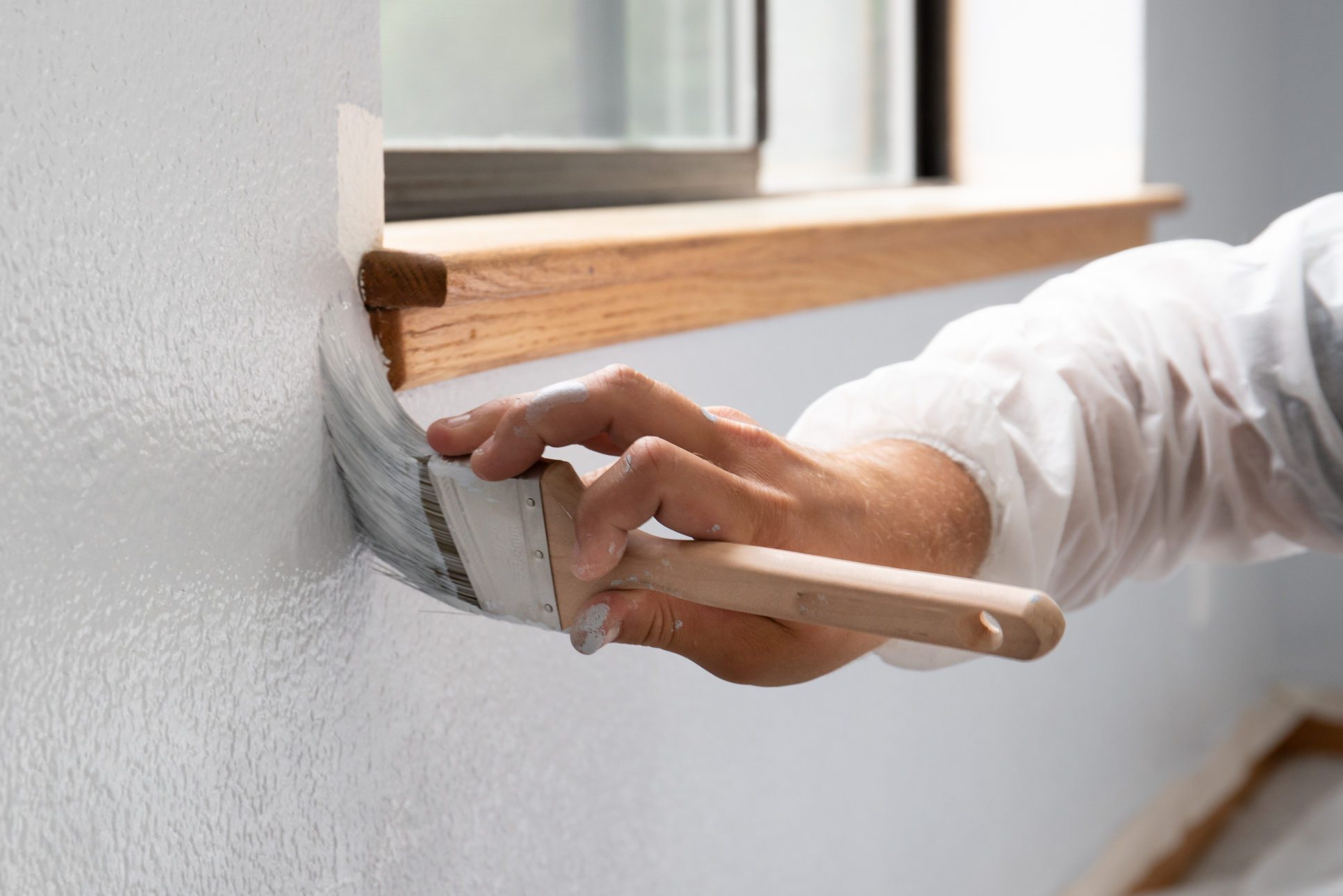 A hand paints a white wall with a brush near a window, wearing a protective sleeve in a well-lit room.