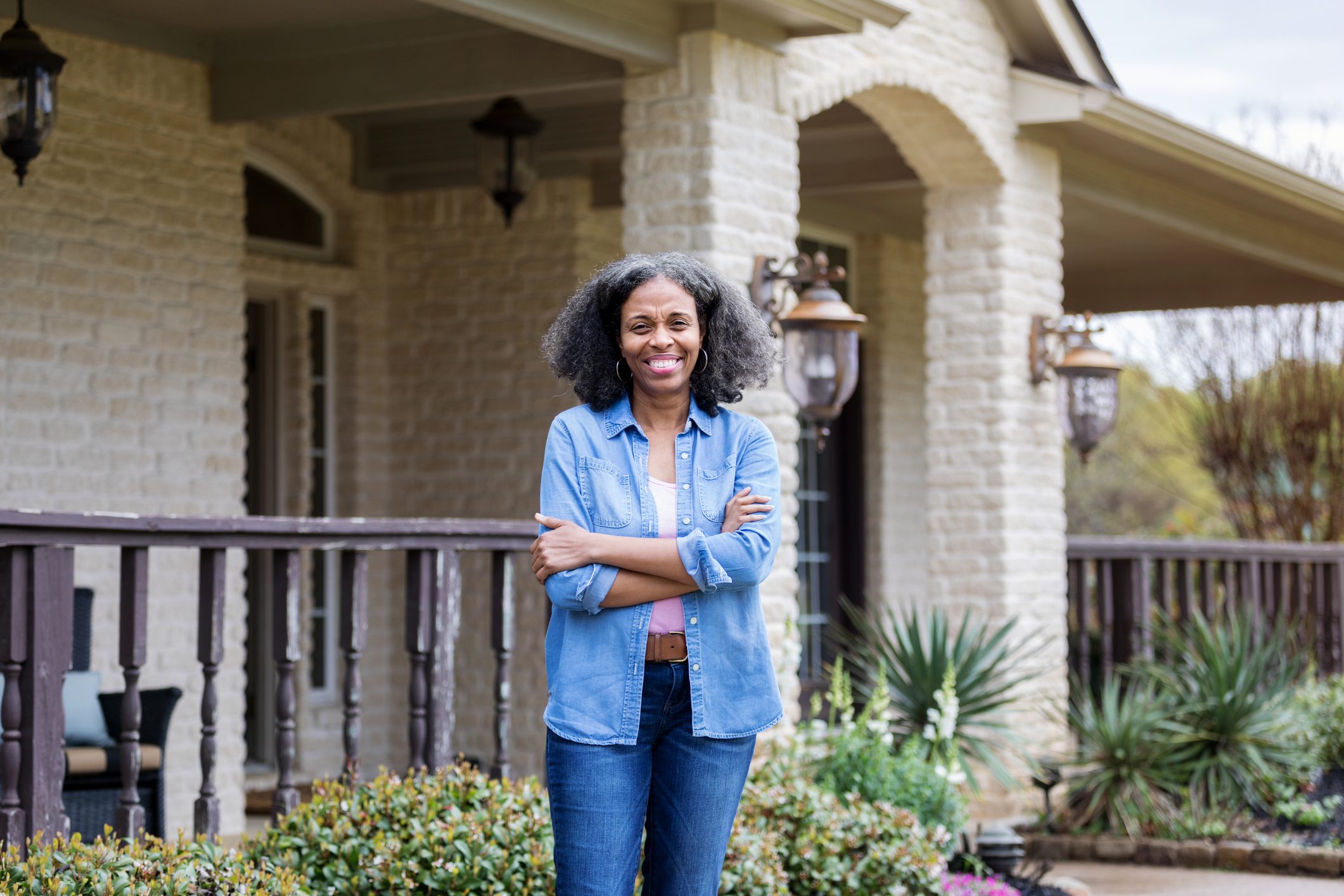 A person stands smiling, arms crossed, on a porch with brick arches, plants, and outdoor lanterns in the background.