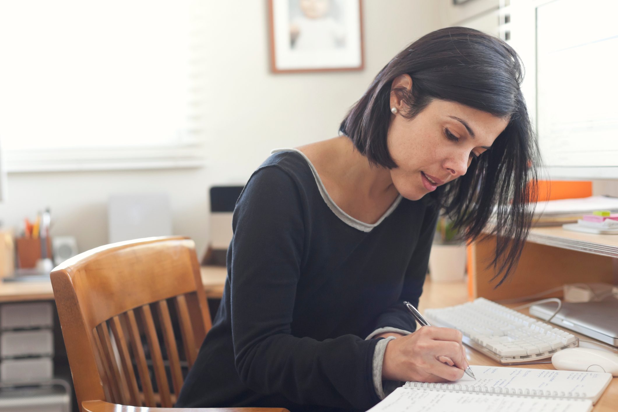 Woman writing in a notebook, focused at a desk with computer and office supplies in a brightly lit room.