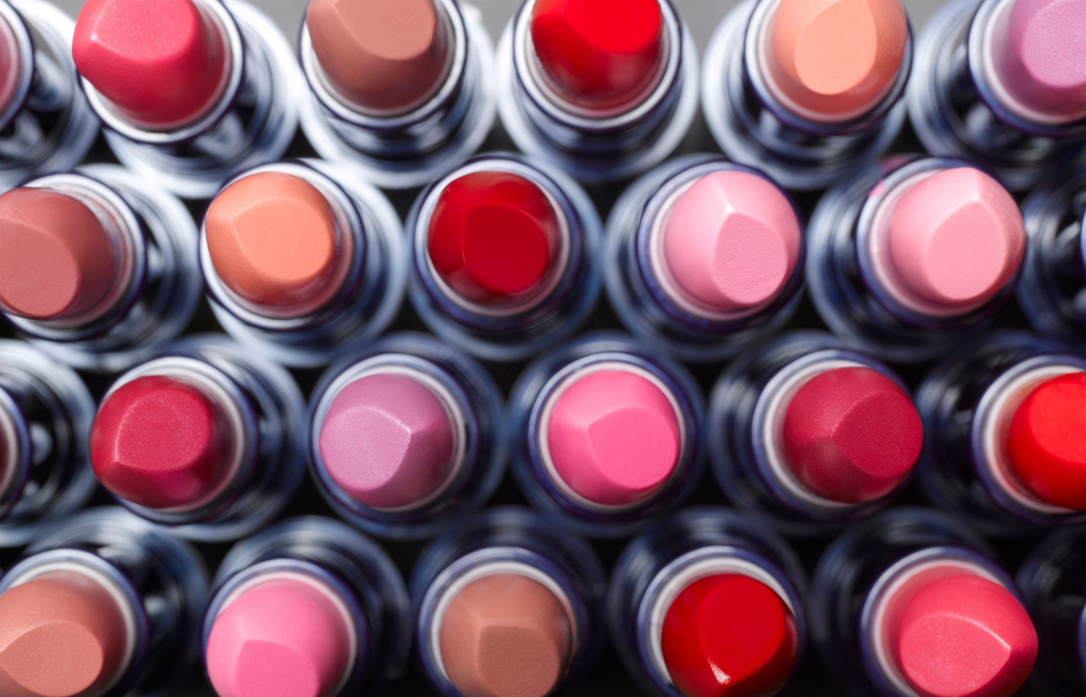 Lipsticks arranged in rows show varied shades, viewed from above, creating a colorful, organized pattern on a neutral background.