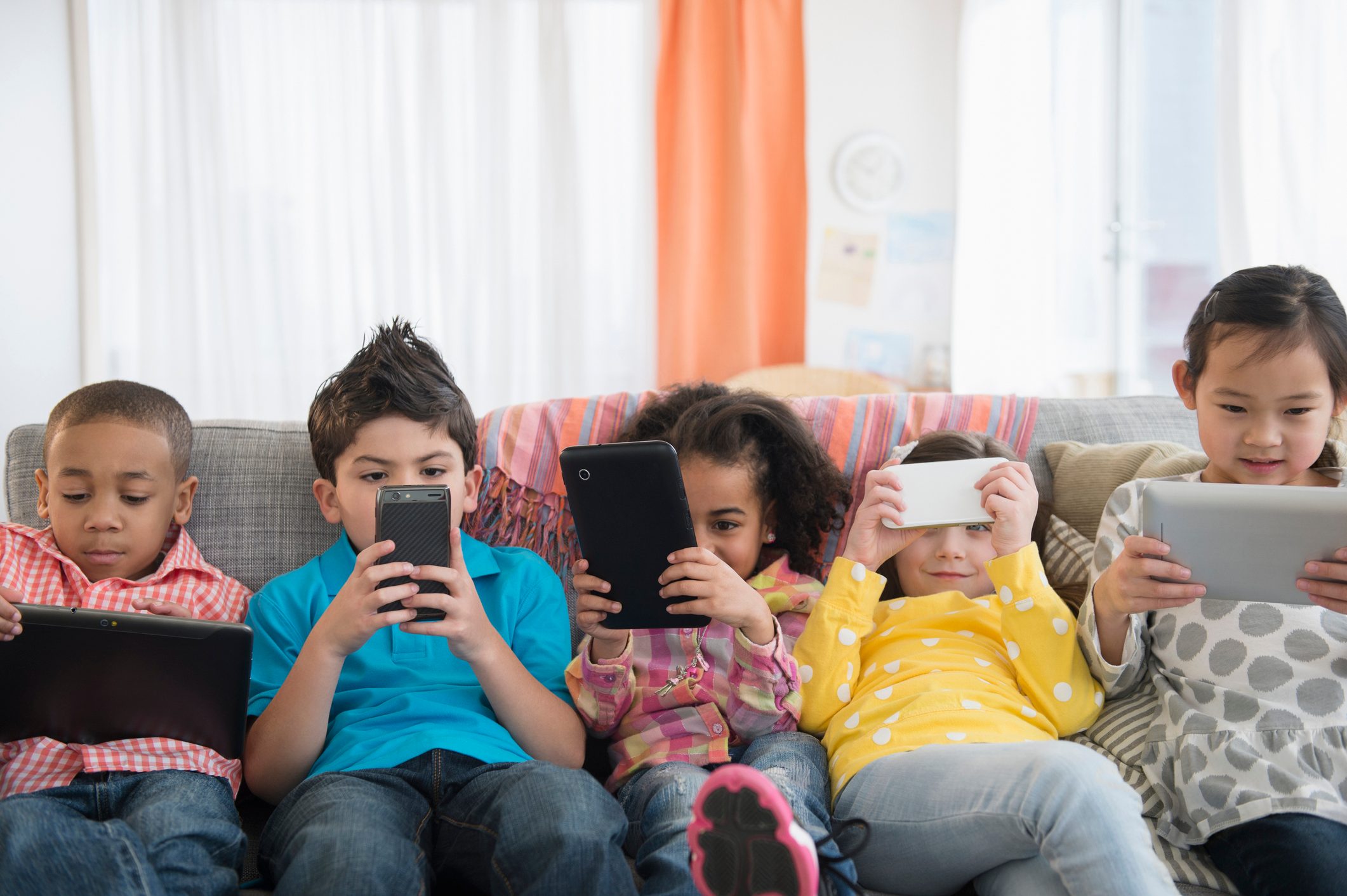 Children gaze at electronic devices while sitting closely on a couch, surrounded by colorful cushions and drapes in a brightly lit room.