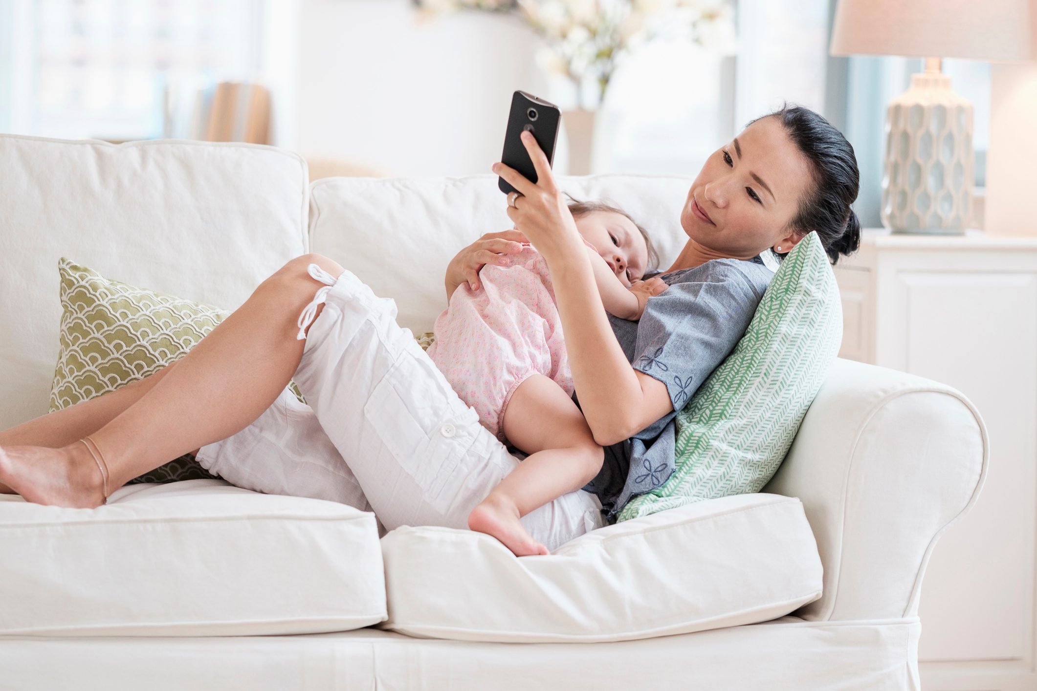 A woman lounges on a white couch, holding a child, while looking at a smartphone in a bright, cozy living room.