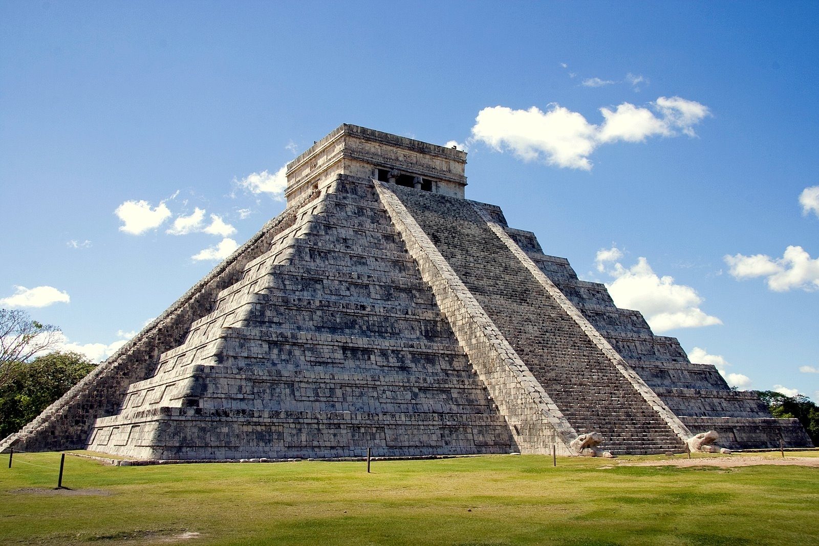 A stone pyramid stands under a clear blue sky, surrounded by green grass, with steps leading to a small temple on top.
