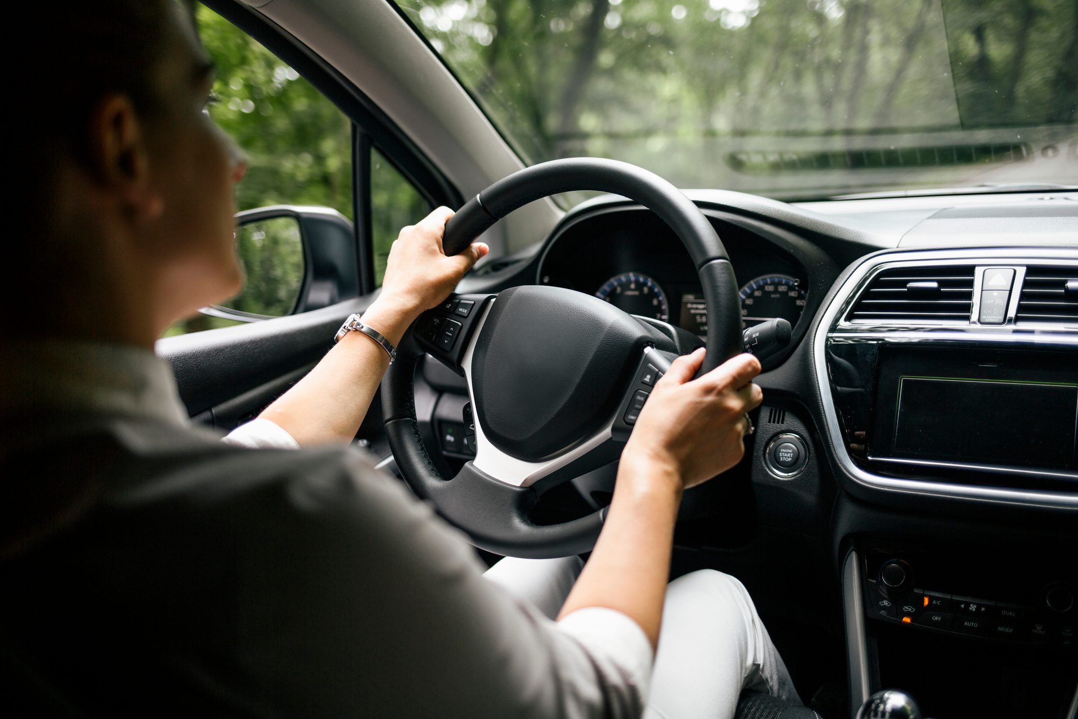 Person drives a car through a wooded area, hands on the steering wheel, dashboard and side mirror visible.