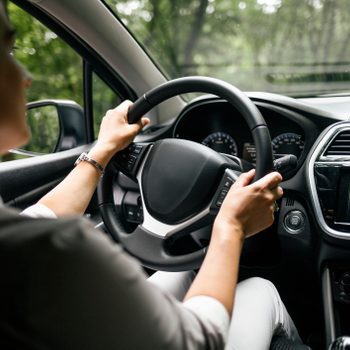 Person drives a car through a wooded area, hands on the steering wheel, dashboard and side mirror visible.