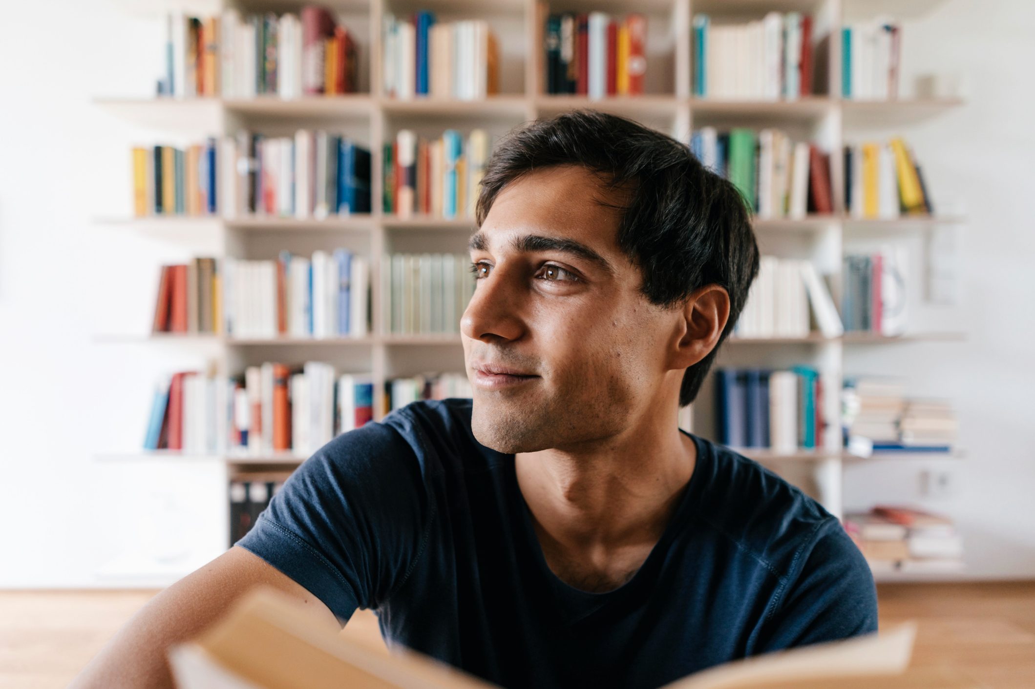 Person reading, smiling, surrounded by a colorful bookshelf in a bright room.