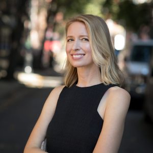 Woman smiling, standing with arms crossed, wearing a sleeveless black dress. Blurred city street with trees and buildings in the background.
