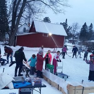 People socialize, skate, and play hockey on a snowy outdoor rink near a red barn, surrounded by trees and houses in the background.